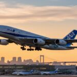 ANAの旅客機が滑走路から離陸する瞬間。夕焼け空と都市のシルエット、遠くにはレインボーブリッジが見える。